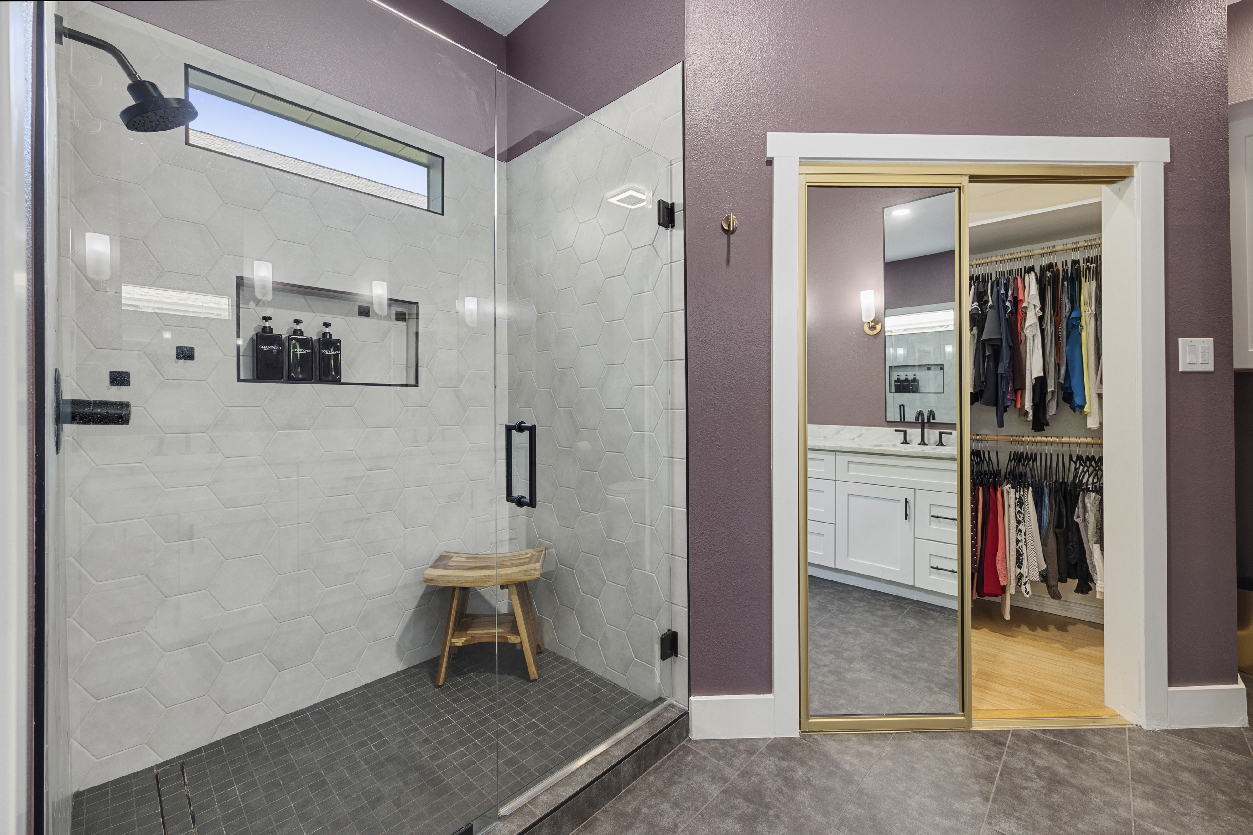 Modern master bathroom with a large walk-in glass shower with hexagon wall tiles, white double vanity cabinets, granite countertops, dark purple walls, and designer black fixtures.