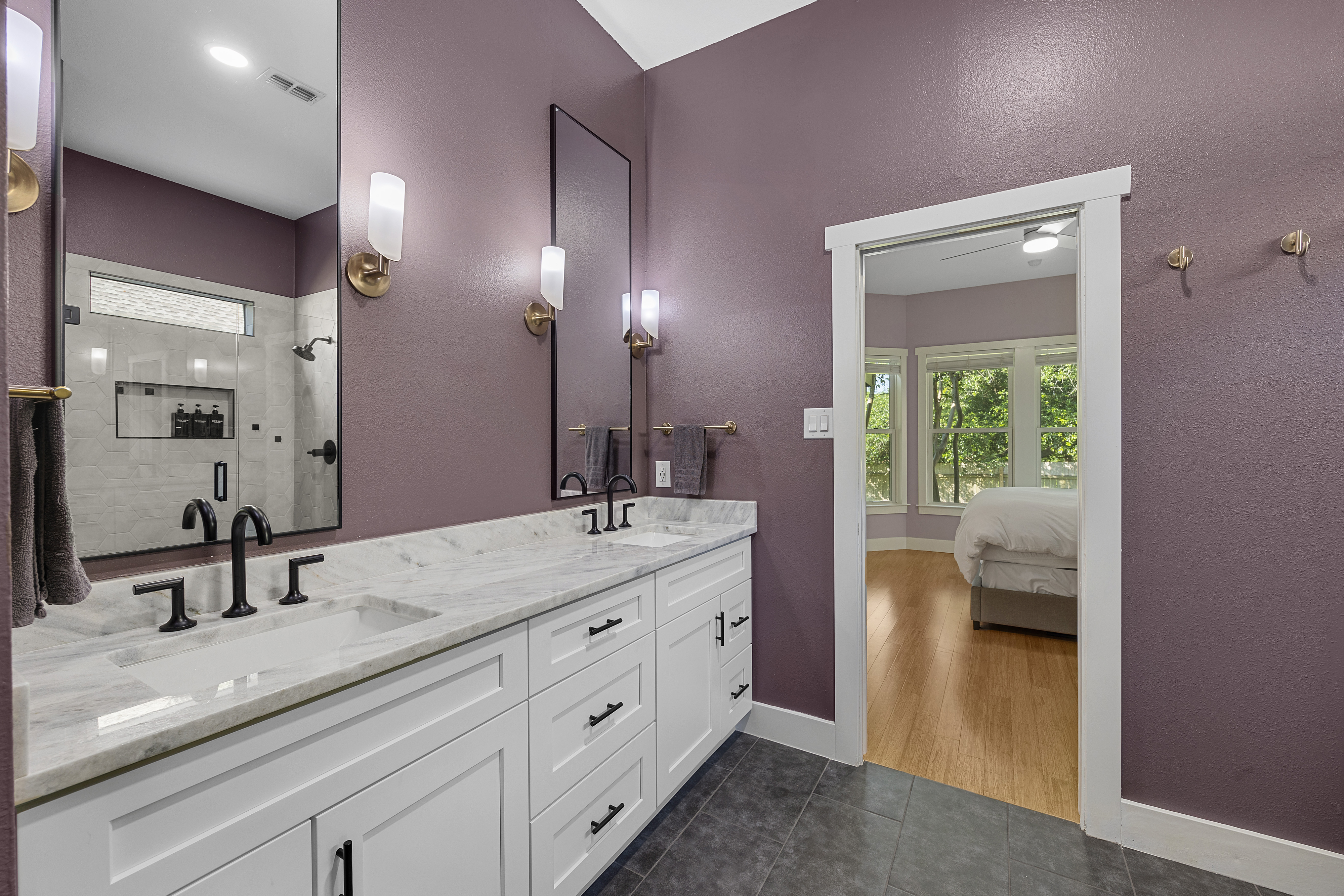 Modern master bathroom with a large walk-in glass shower with hexagon wall tiles, white double vanity cabinets, granite countertops, dark purple walls, and designer black fixtures.