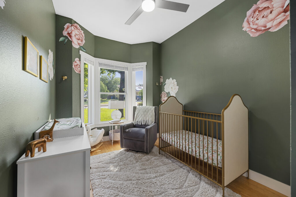 Baby girl's nursery with green, pink, and gold decor, featuring a crib, changing table, dresser, rocking chair, white rug, and tall ceilings.