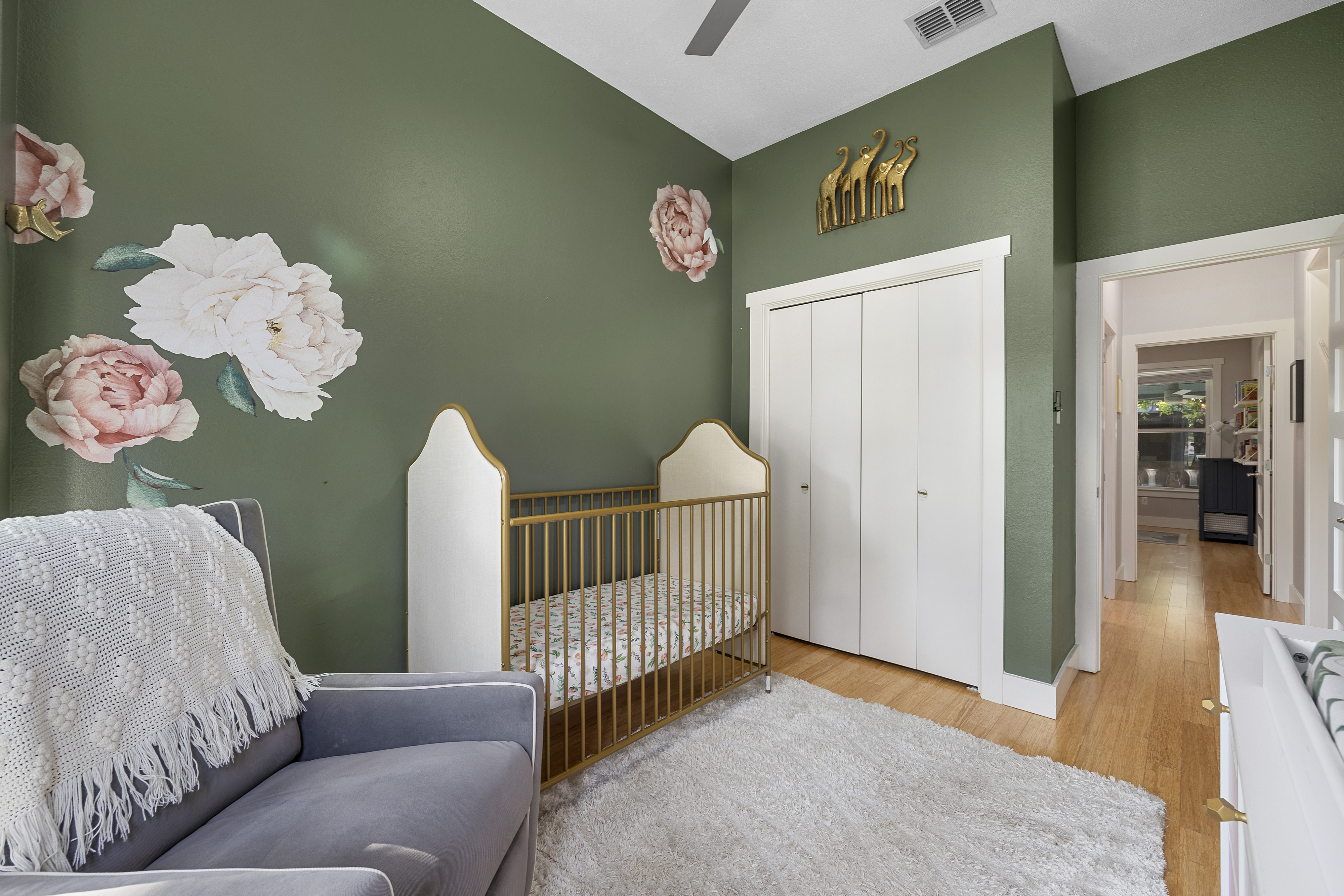 Baby girl's nursery with green, pink, and gold decor, featuring a crib, changing table, dresser, rocking chair, white rug, and tall ceilings.